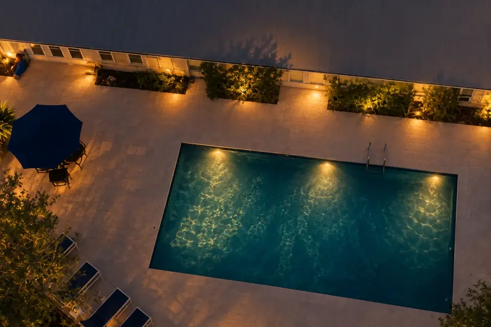 The swimming pool from above at night, lit from within, loungers and a parasol along the warm-lit deck.