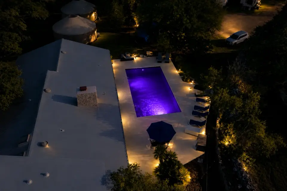 The pool terrace from the air after dark, the water lit violet beside the wellness centre roofline.