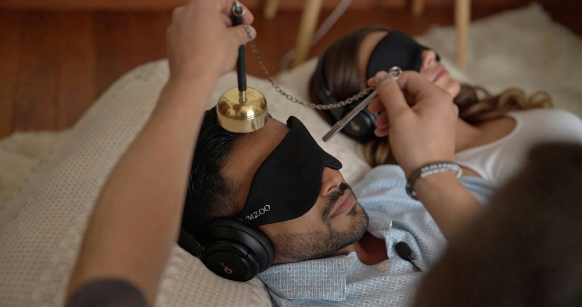 A guide sounding a brass bowl over two people resting in eye masks and headphones during a ceremony